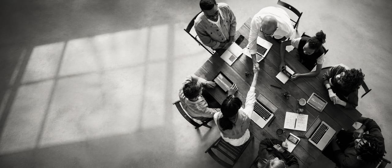 Team am Besprechungstisch von oben, Handschlag über Laptops und Notizen — AHEAD OF TIME